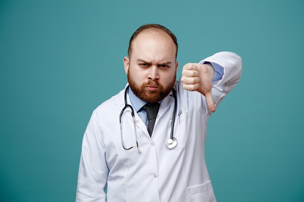unpleased young male doctor wearing medical coat and stethoscope around his neck looking at camera showing thumb down isolated on blue background