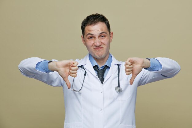 недовольный молодой врач-мужчина в медицинском халате и стетоскопе на шее смотрит в камеру, показывая большие пальцы вниз на оливково-зеленом фоне