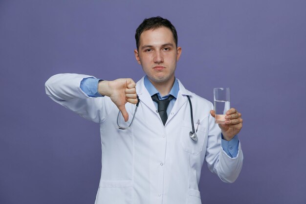 Unpleased young male doctor wearing medical robe and stethoscope around neck showing glass of water looking at camera showing thumb down isolated on purple background