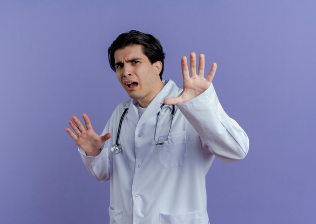 Unpleased young male doctor wearing medical robe and stethoscope  doing no gesture isolated on purple wall with copy space