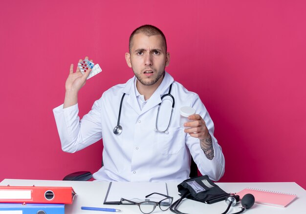 Unpleased young male doctor wearing medical robe and stethoscope sitting at desk with work tools holding medical beaker and packs of tablets and capsules isolated on pink background