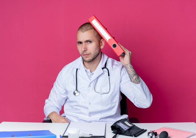 Unpleased young male doctor wearing medical robe and stethoscope sitting at desk with work tools touching head with folder isolated on pink background