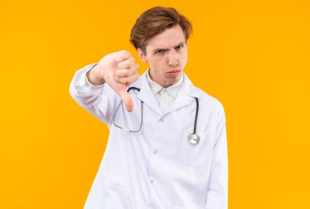 Un giovane medico maschio scontento che indossa una tunica medica con uno stetoscopio che mostra il pollice verso il basso isolato sulla parete arancione
