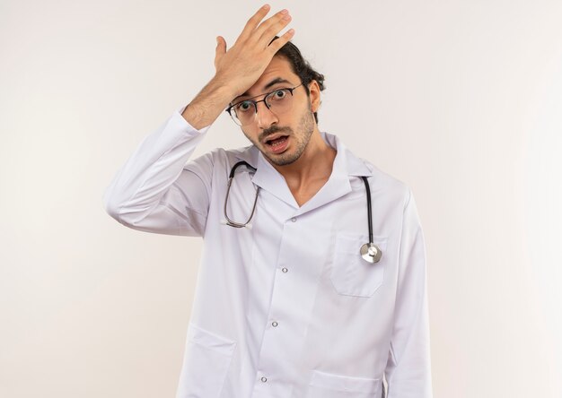 Unpleased young male doctor with optical glasses wearing white robe with stethoscope putting hand on forehead on isolated white wall with copy space