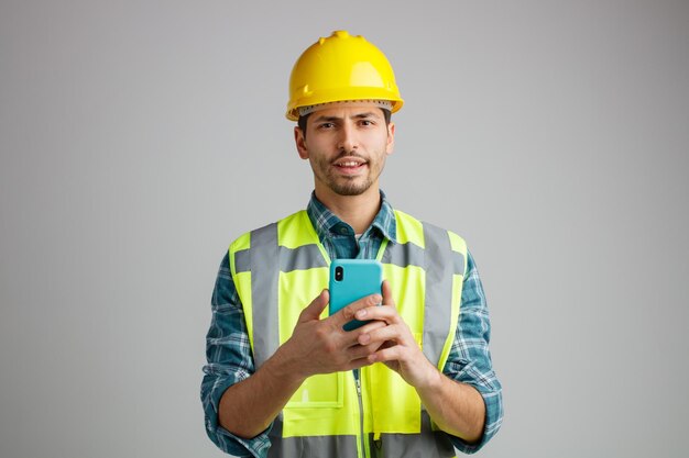 Unpleased young male engineer wearing safety helmet and uniform holding mobile phone with both hands looking at camera isolated on white background