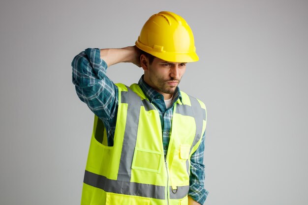 Unpleased young male engineer wearing safety helmet and uniform looking at side keeping hand on back of head having headache isolated on white background