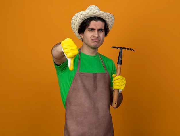 Бесплатное фото Недовольный молодой мужчина-садовник в униформе в садовой шляпе с перчатками, держащими грабли, показывая большой палец вниз, изолированный на оранжевом
