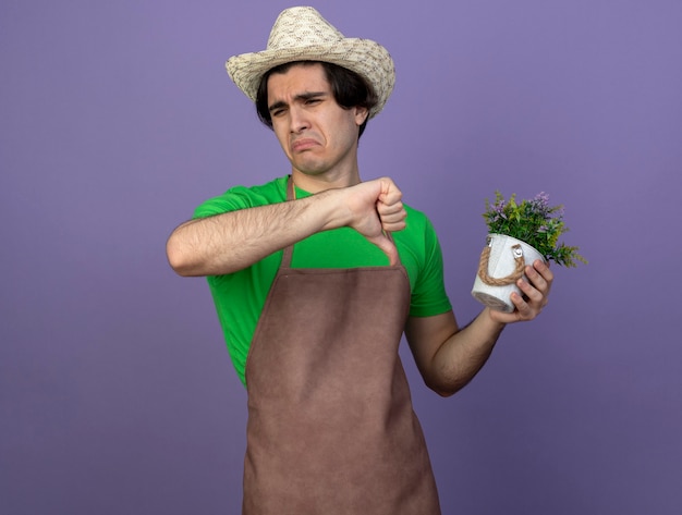 Unpleased young male gardener in uniform wearing gardening hat holding flower in flowerpot showing thumb down