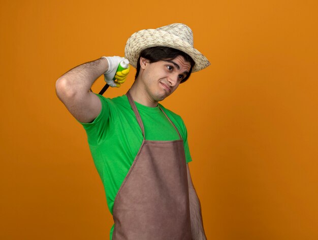 Unpleased young male gardener in uniform wearing gardening hat with gloves scratching with rake shoulder isolated on orange