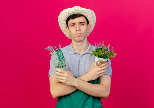 Foto gratuita un giovane giardiniere maschio scontento che indossa un cappello da giardinaggio incrocia le braccia tenendo i vasi da fiori