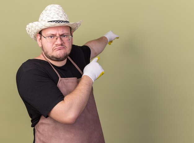 Unpleased young male gardener wearing gardening hat and gloves points at behind isolated on olive green wall with copy space