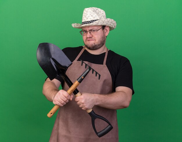 Unpleased young male gardener wearing gardening hat holding and crossing spade with rake isolated on green wall