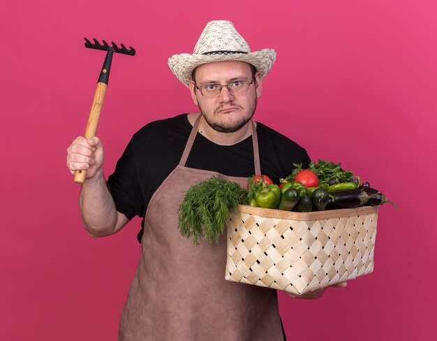 분홍색 벽에 고립 된 갈퀴를 올리는 야채 바구니를 들고 원예 모자를 쓰고 불쾌한 젊은 남성 정원사