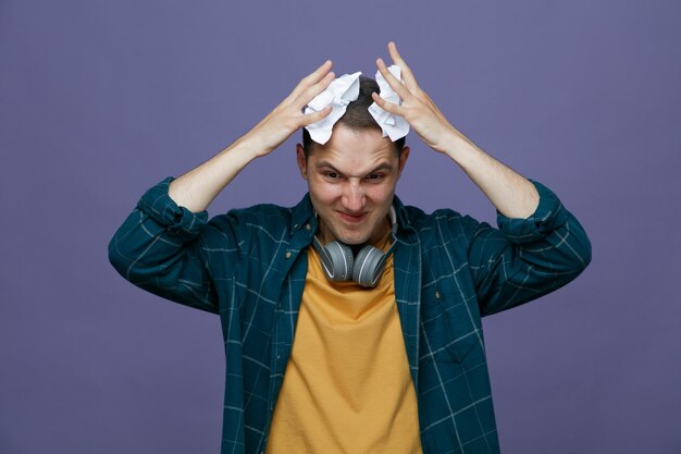 unpleased young male student wearing headphones around neck putting torn and crushed pieces of exam papers on head looking down isolated on purple background