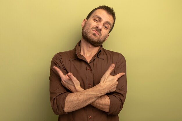 Unpleased young man keeping hands crossed pointing at sides isolated on olive green wall