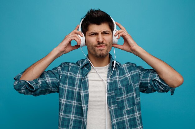 Unpleased young man wearing headphones looking at camera grabbing headphones isolated on blue background