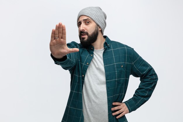 Unpleased young man wearing winter hat keeping hand on waist looking at camera showing stop gesture isolated on white background