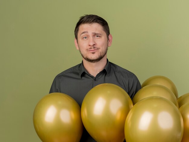 Unpleased young party guy wearing black shirt standing among balloons isolated on olive green