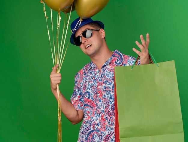 Ragazzo di partito giovane insoddisfatto che indossa cappello blu e bicchieri in possesso di palloncini con borsa regalo e diffusione di mano isolata sul verde