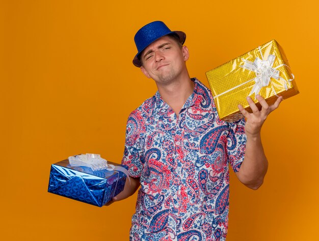 Unpleased young party guy wearing blue hat holding gift boxes isolated on orange