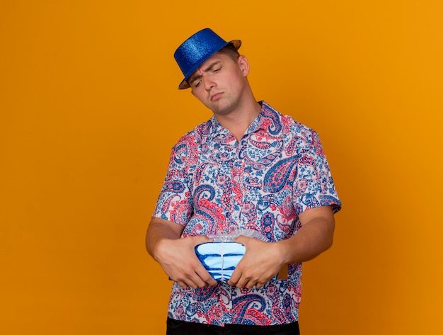 Unpleased young party guy wearing blue hat holding and looking at gift box isolated on orange