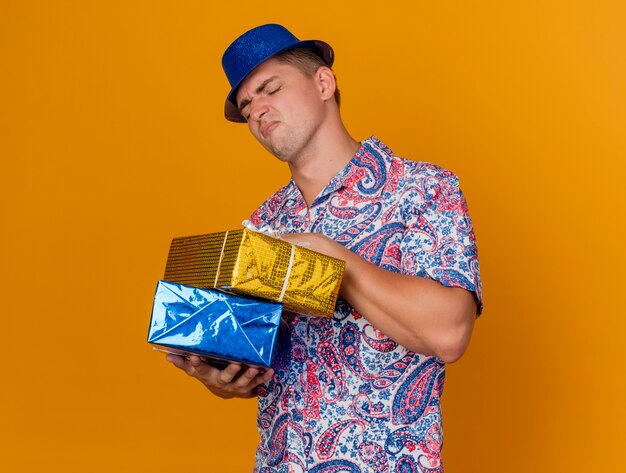 Unpleased young party guy wearing blue hat holding and looking at gift boxes isolated on orange