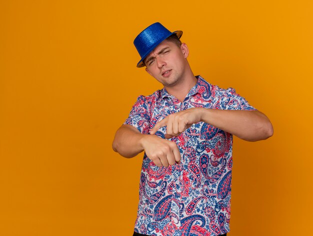 Unpleased young party guy wearing blue hat showing wrist clock gesture isolated on orange