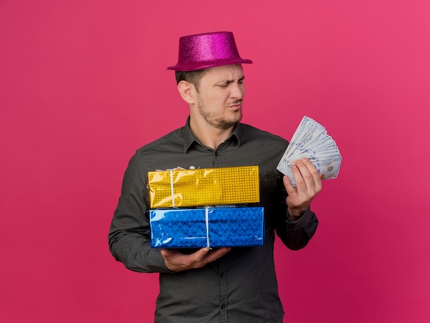 Foto gratuita ragazzo di partito giovane insoddisfatto che indossa il cappello rosa tenendo scatole regalo e guardando i contanti in mano isolato sul rosa