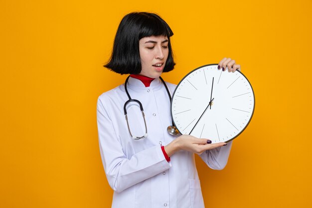 Unpleased young pretty caucasian girl in doctor uniform with stethoscope holding and looking at clock