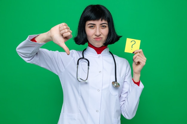 Unpleased young pretty caucasian girl in doctor uniform with stethoscope thumbing down and holding question note 