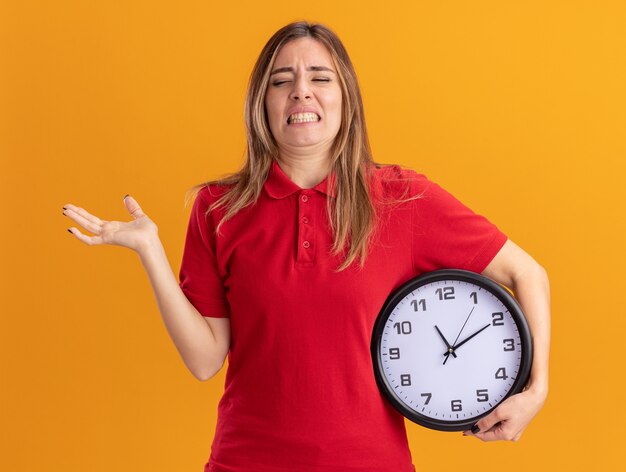 Unpleased young pretty caucasian girl holds clock and keeps hand open on orange