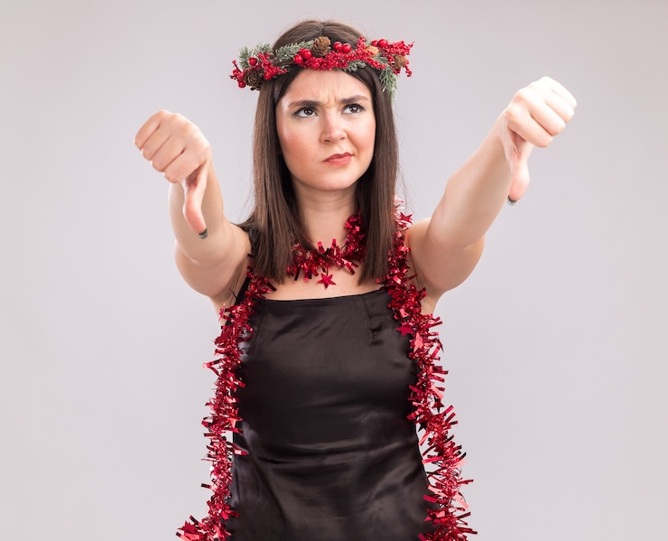 मुफ़्त फोटो unpleased young pretty caucasian girl wearing christmas head wreath and tinsel garland around neck looking up showing thumbs down isolated on white background