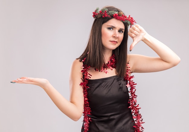 Unpleased young pretty caucasian girl wearing christmas head wreath and tinsel garland around neck showing empty hand and thumb down looking at camera isolated on white background