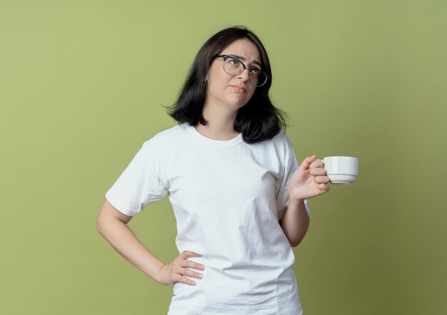 Unpleased young pretty caucasian girl wearing glasses looking up putting hand on waist holding cup isolated on olive green background with copy space