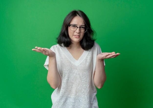 Unpleased young pretty caucasian girl wearing glasses showing empty hands isolated on green background