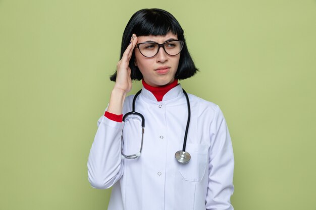 Unpleased young pretty caucasian girl with optical glasses in doctor uniform with stethoscope puts hand on temple 