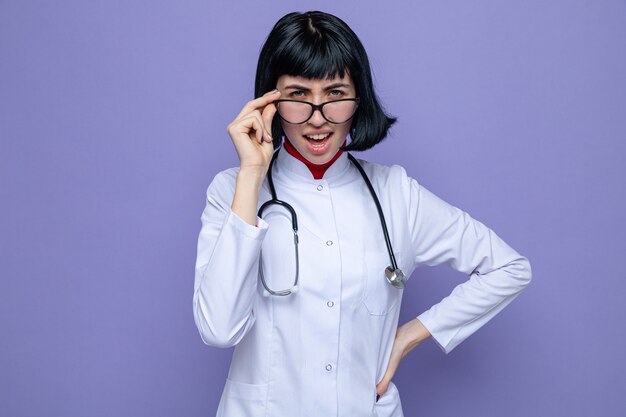 Unpleased young pretty caucasian girl with optical glasses wearing doctor uniform with stethoscope 