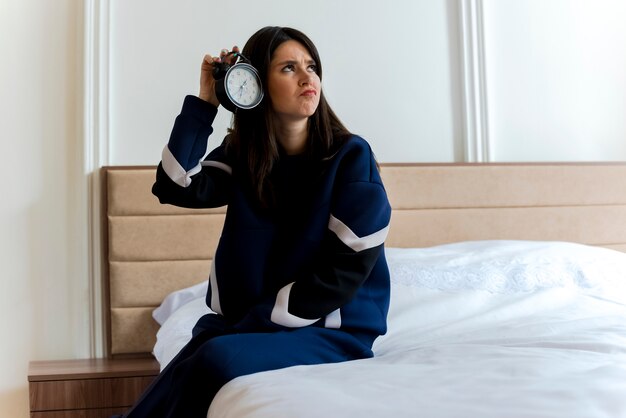 Unpleased young pretty caucasian woman sitting on bed in bedroom holding alarm clock near head looking at side