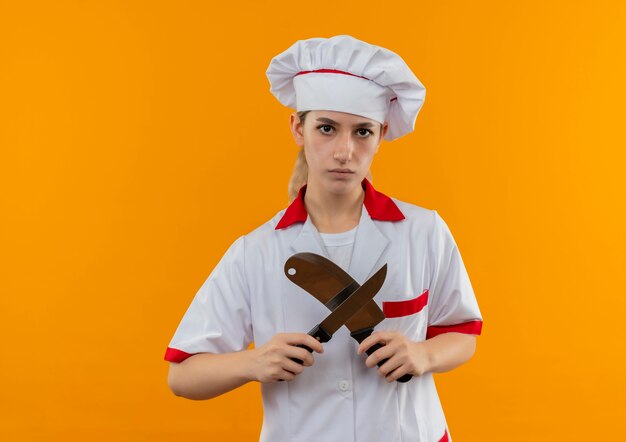 오렌지 공간에 고립 된 칼과 칼로 몸짓을하지 않는 요리사 유니폼에 불쾌한 젊은 예쁜 요리사