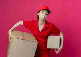 Бесплатное фото Недовольная молодая симпатичная доставщица в красной униформе и кепке смотрит в сторону и держит картонные коробки