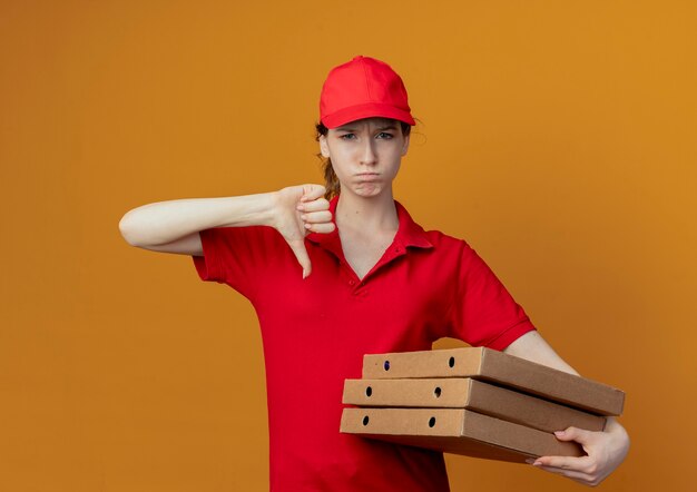 Недовольная молодая симпатичная доставщица в красной форме и кепке держит пакеты с пиццей и показывает большой палец вниз, изолированную на оранжевом фоне