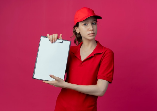 Giovane ragazza graziosa di consegna poco soddisfatta in uniforme rossa e cappuccio che mostra appunti isolati su fondo cremisi con lo spazio della copia