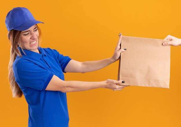 Unpleased young pretty delivery girl in uniform gives paper package to someone on orange