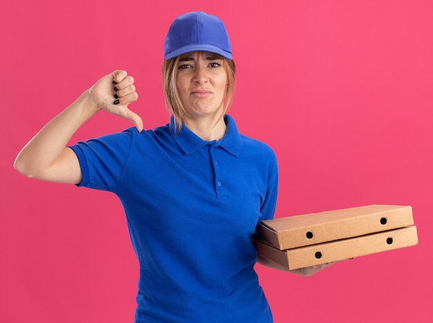 Недовольная молодая симпатичная доставщица в униформе держит коробки с пиццей и большими пальцами смотрит на розовый