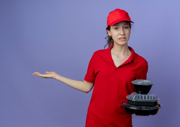 Unpleased young pretty delivery girl wearing red uniform and cap holding food containers and showing empty hand isolated on purple background with copy space