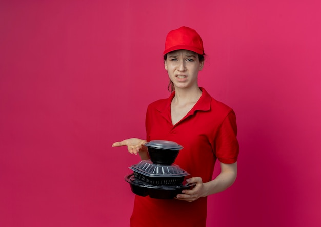 빨간 유니폼과 모자를 들고 복사 공간이 진홍색 배경에 고립 된 식품 용기에서 손으로 가리키는 불쾌한 젊은 예쁜 배달 소녀