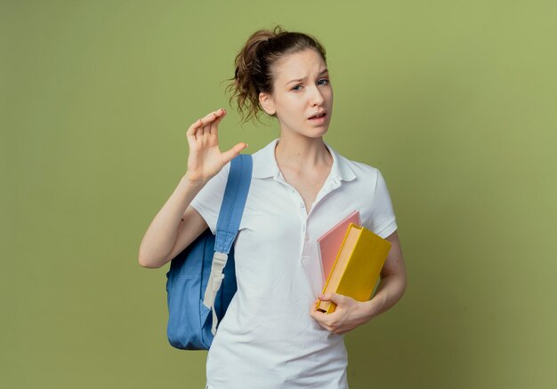 Unpleased young pretty female student wearing back bag holding book and note pad showing size isolated on olive green background with copy space