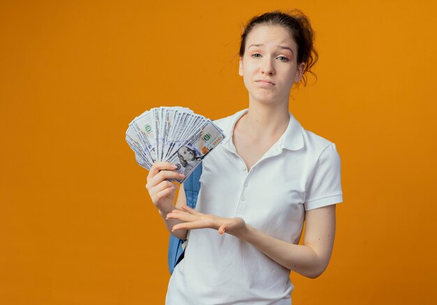 Недовольная молодая симпатичная студентка в задней сумке, держащая и указывающая рукой на деньги, изолированные на оранжевом фоне с копией пространства