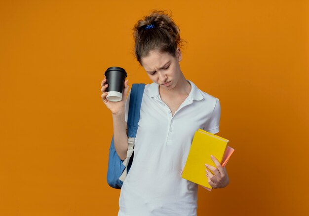 コピースペースでオレンジ色の背景に分離された本のメモ帳ペンとプラスチック製のコーヒーカップを保持して見下ろしているバックバッグを身に着けている不機嫌な若いきれいな女性