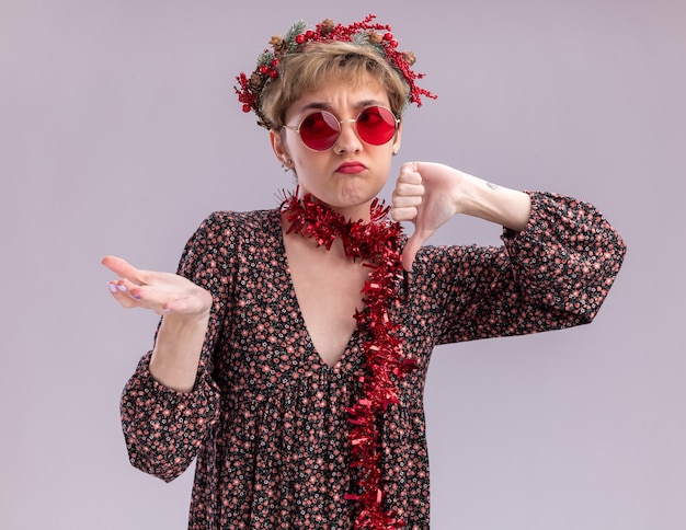 Free photo unpleased young pretty girl wearing christmas head wreath and tinsel garland around neck with glasses looking at side showing empty hand isolated on white wall